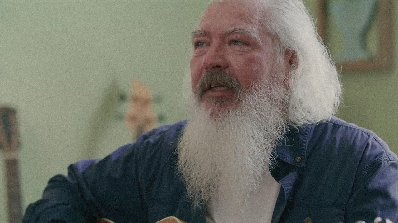 Gary L. Clark, with his long white beard, seated with guitars on the wall behind him