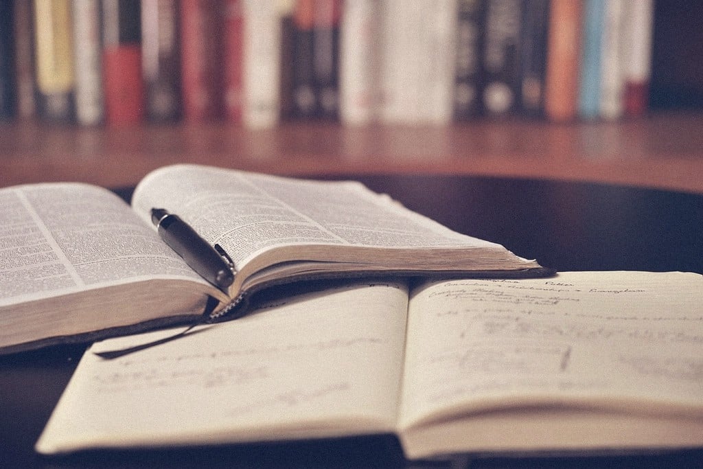 Two open books and a pen on a wooden desk, bookshelves softly out of focus behind