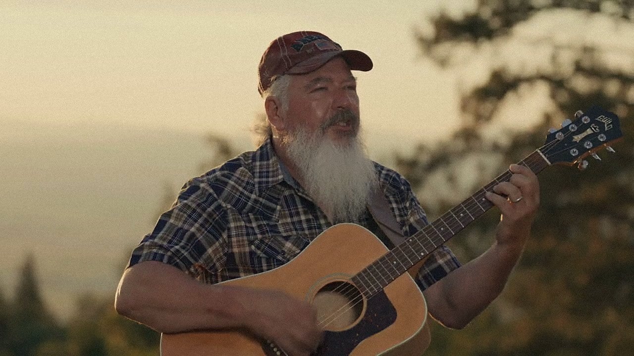 Gary L. Clark playing an acoustic guitar outdoors at golden hour, from the music video for She's Red White and Blue